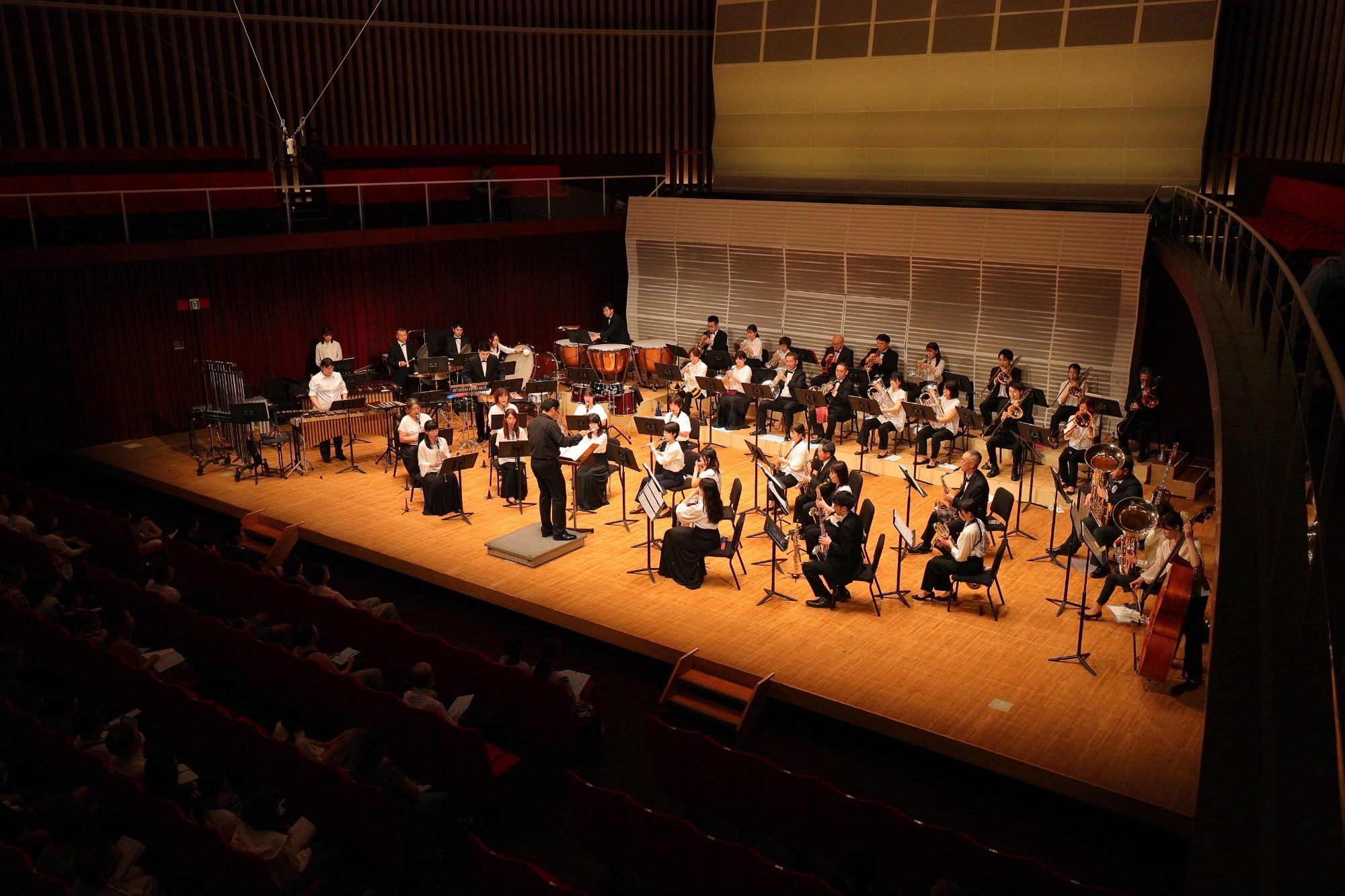 Wind Orchestra Concert Performance コンサートホールの舞台で演奏する吹奏楽団の様子。黒と白の衣装を着た奏者たちが指揮者の指示のもと演奏している